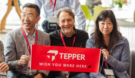 Three Tepper School of Business alumni, two men and one woman, holding a red sign that says 'Tepper, Wish you were here.' at Reunion Weekend